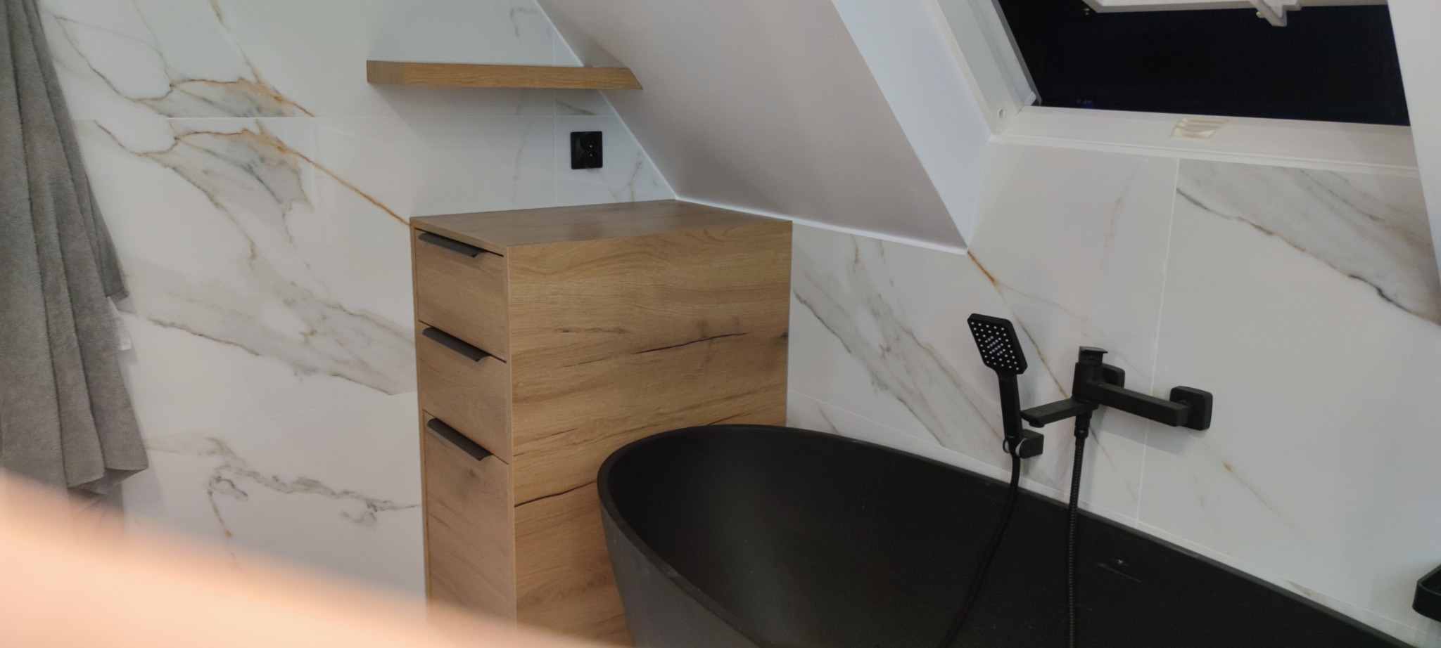 Drewniana szafka z szufladami obok czarnej wanny w łazience na poddaszu. Białe płytki imitujące marmur na ścianach, drewniana półka.