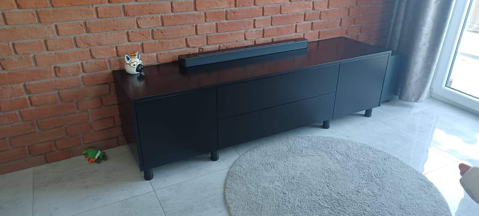 Nowoczesna, czarna szafka RTV z szufladami na tle ceglanej ściany. Na blacie stoi głośnik i ceramiczna figurka psa w okularach. U dołu widoczny okrągły dywan.