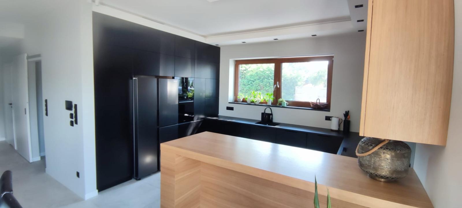 Nowoczesna kuchnia: czarne szafki, drewniany blat, okno z roślinami. Minimalistyczny design z akcentem naturalnego drewna i świeżej zieleni. Funkcjonalność i estetyka.