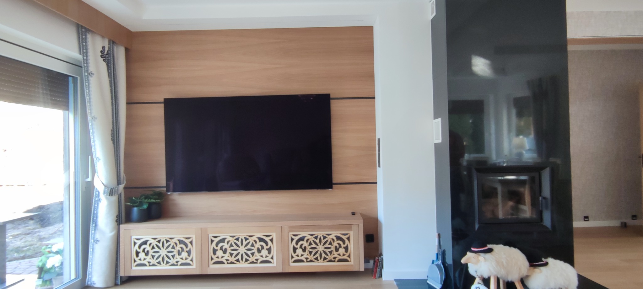 Drewniana ściana TV z szafką RTV o ażurowych frontach, obok kominek w czarnej obudowie. Okno z zasłoną po lewej stronie. Jasne, naturalne wnętrze.