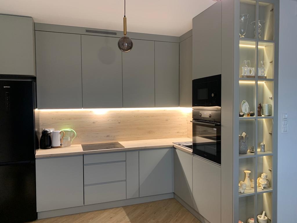 Nowoczesna kuchnia w odcieniach szarości z oświetleniem LED, zabudową AGD i przeszkloną witryną. Minimalistyczny design, drewniany blat, czarna lodówka.