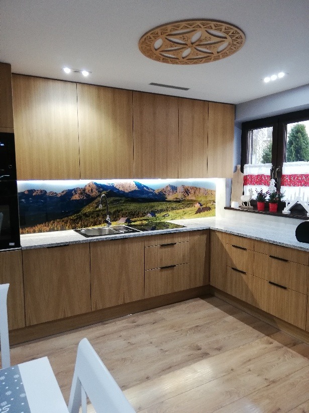 Jasna kuchnia z drewnianymi frontami i blatem w odcieniach szarości. Nad blatem panel z motywem górskim. Na suficie dekoracyjny, okrągły element oświetleniowy.