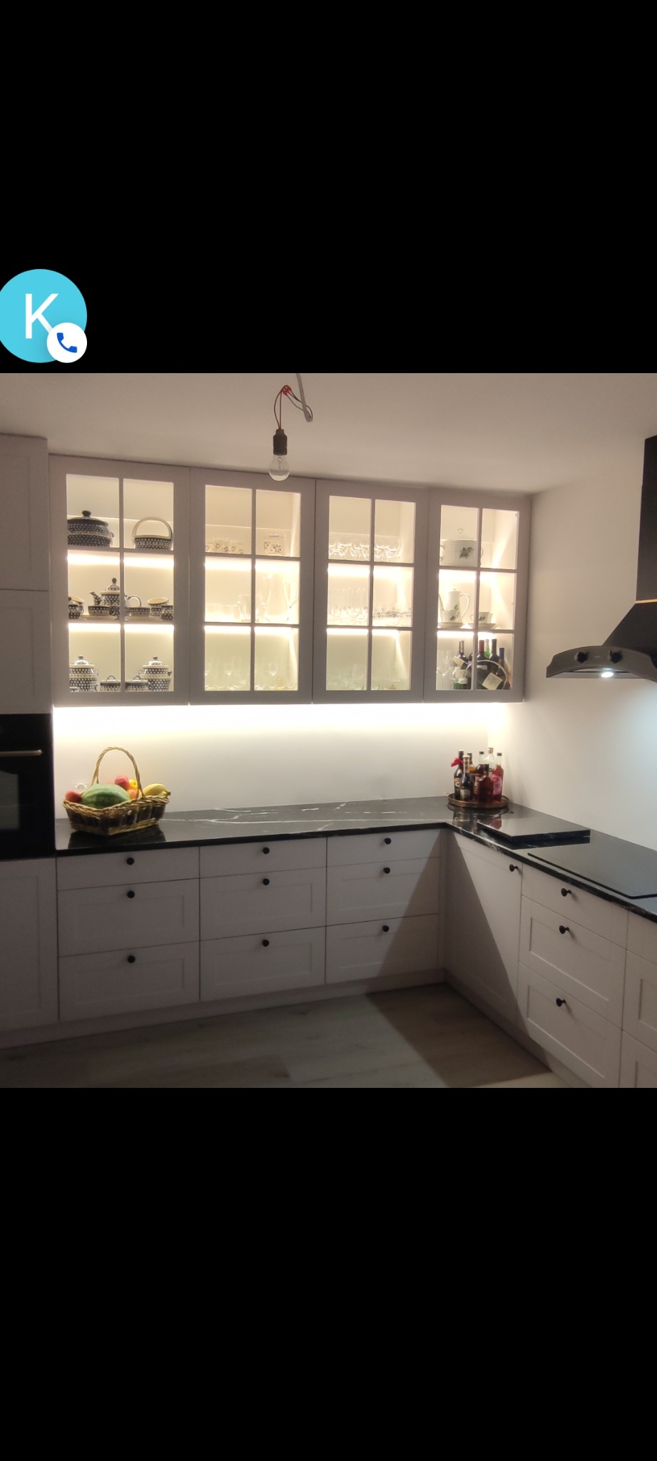 Jasna kuchnia z czarnym blatem, oświetleniem LED pod szafkami i przeszklonymi szafkami wiszącymi wypełnionymi naczyniami i dekoracjami. Widoczny okap.