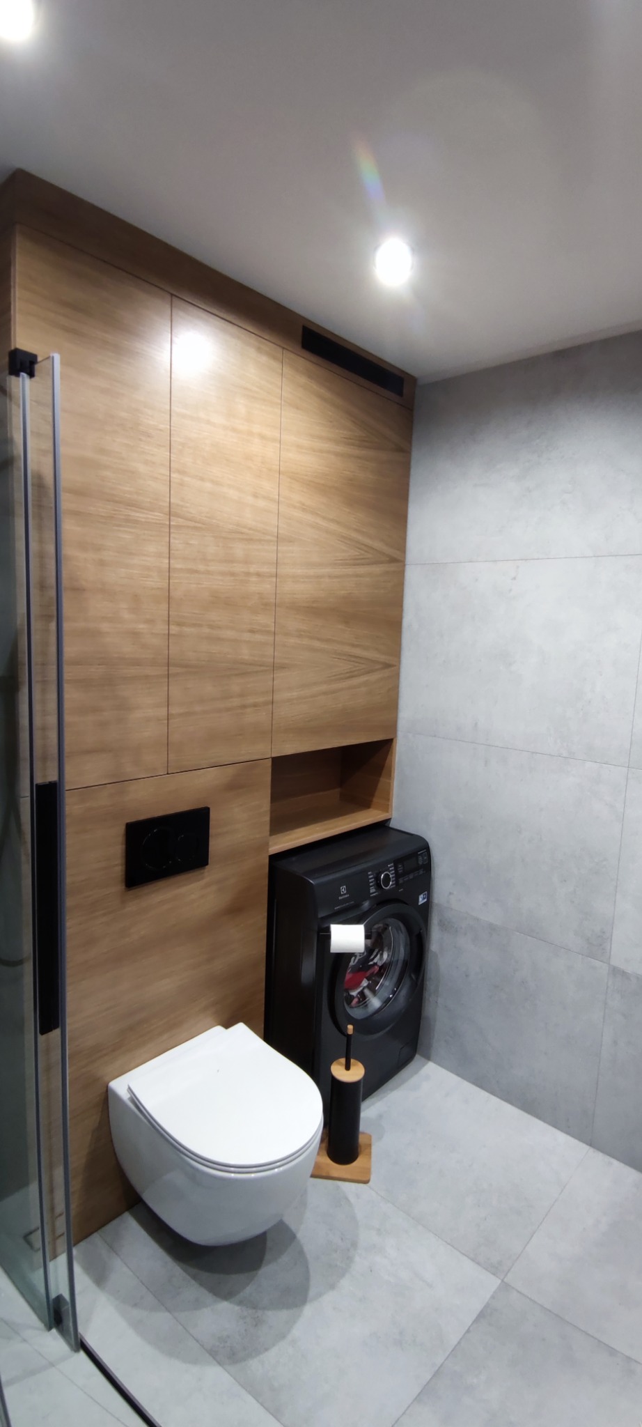 Nowoczesna łazienka z drewnianą zabudową meblową, podwieszaną toaletą i czarną pralką. Minimalistyczny design z szarymi płytkami.