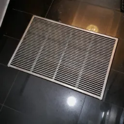 Metalowa wycieraczka kratowa z drobnymi szczelinami, ułożona na ciemnych, błyszczących płytkach podłogowych, widoczna z góry pod kątem.