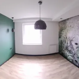 Panoramiczny widok na odnowione wnętrze z zieloną ścianą akcentową z pionowymi złotymi paskami, fototapetą z motywem roślinnym na drugiej ścianie i drewnianą podłogą.