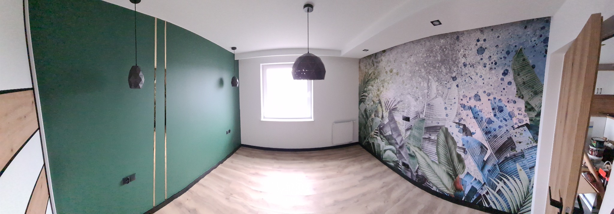 Panoramiczny widok na odnowione wnętrze z zieloną ścianą akcentową z pionowymi złotymi paskami, fototapetą z motywem roślinnym na drugiej ścianie i drewnianą podłogą.