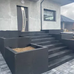 Nowoczesne, ciemnoszare schody zewnętrzne z kostki brukowej prowadzące do drzwi wejściowych domu z szarą elewacją, widoczne donice na rośliny z ziemią i fragment podjazdu z kostki brukowej...