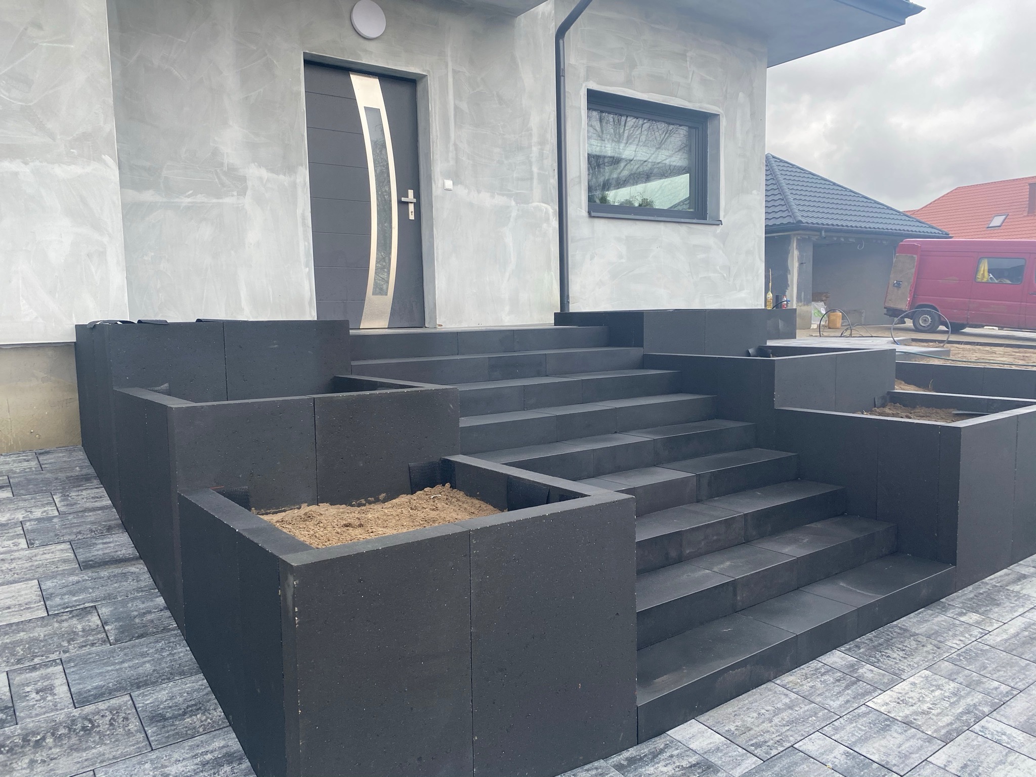 Nowoczesne, ciemnoszare schody zewnętrzne z kostki brukowej prowadzące do drzwi wejściowych domu z szarą elewacją, widoczne donice na rośliny z ziemią i fragment podjazdu z kostki brukowej...