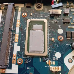 Otwarta obudowa laptopa z widoczną płytą główną i nałożoną pastą termoprzewodzącą na procesorze, widoczne elementy elektroniczne i złącza.