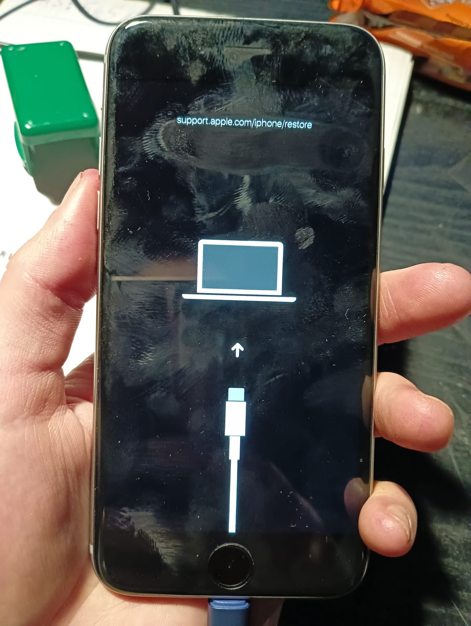 Czarny smartfon w dłoni, ekran wyświetla ikonę laptopa, strzałkę w górę i symbol kabla USB, sugerując proces przywracania systemu z komunikatem 'support.apple.com/iphone/restore'. Ekran jest nieco...