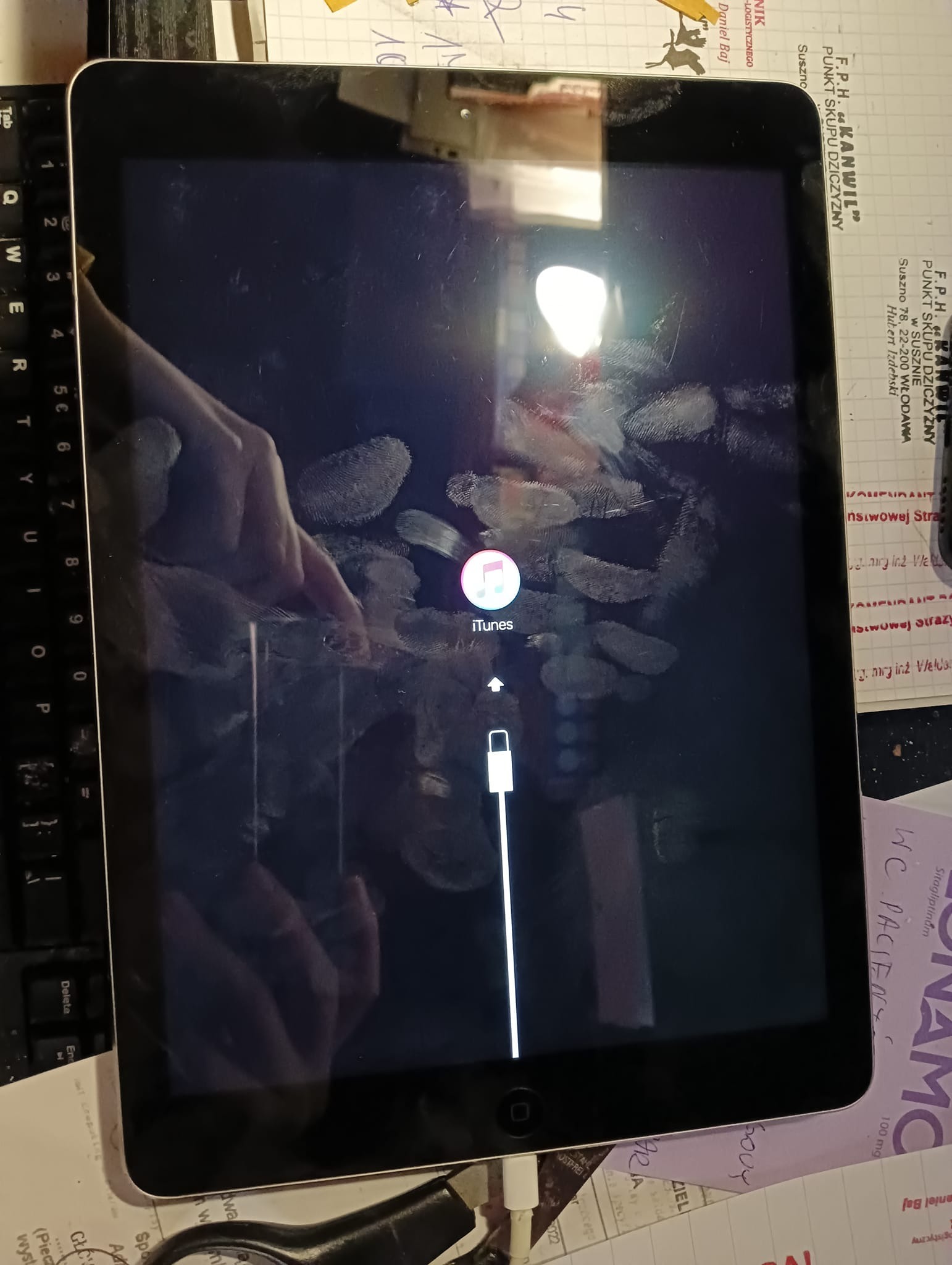 Tablet z widocznym symbolem iTunes i śladami palców na ekranie, podłączony do ładowania, leżący na biurku z dokumentami i klawiaturą.