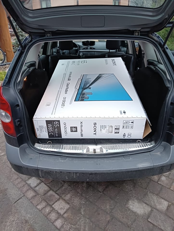 Pudełko z telewizorem Sony Bravia 65 cali w bagażniku samochodu osobowego, widoczne specyfikacje techniczne na opakowaniu.
