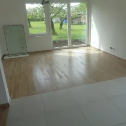 Wnętrze pokoju z dwiema różnymi podłogami: panele imitujące drewno i białe płytki, duże okno z widokiem na ogród, ściany pomalowane na biało, szyba oparta o ścianę.