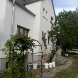 Mężczyzna w kraciastej koszuli maluje białą elewację domu jednorodzinnego, stojąc na drewnianej drabinie, z widocznymi wiadrami z farbą i ozdobną konstrukcją roślinną w ogrodzie.