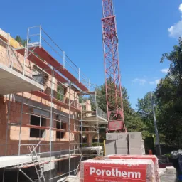 Budowa domu jednorodzinnego z czerwonej cegły Porotherm, widoczne rusztowania, dźwig i palety z materiałem budowlanym na tle błękitnego nieba.