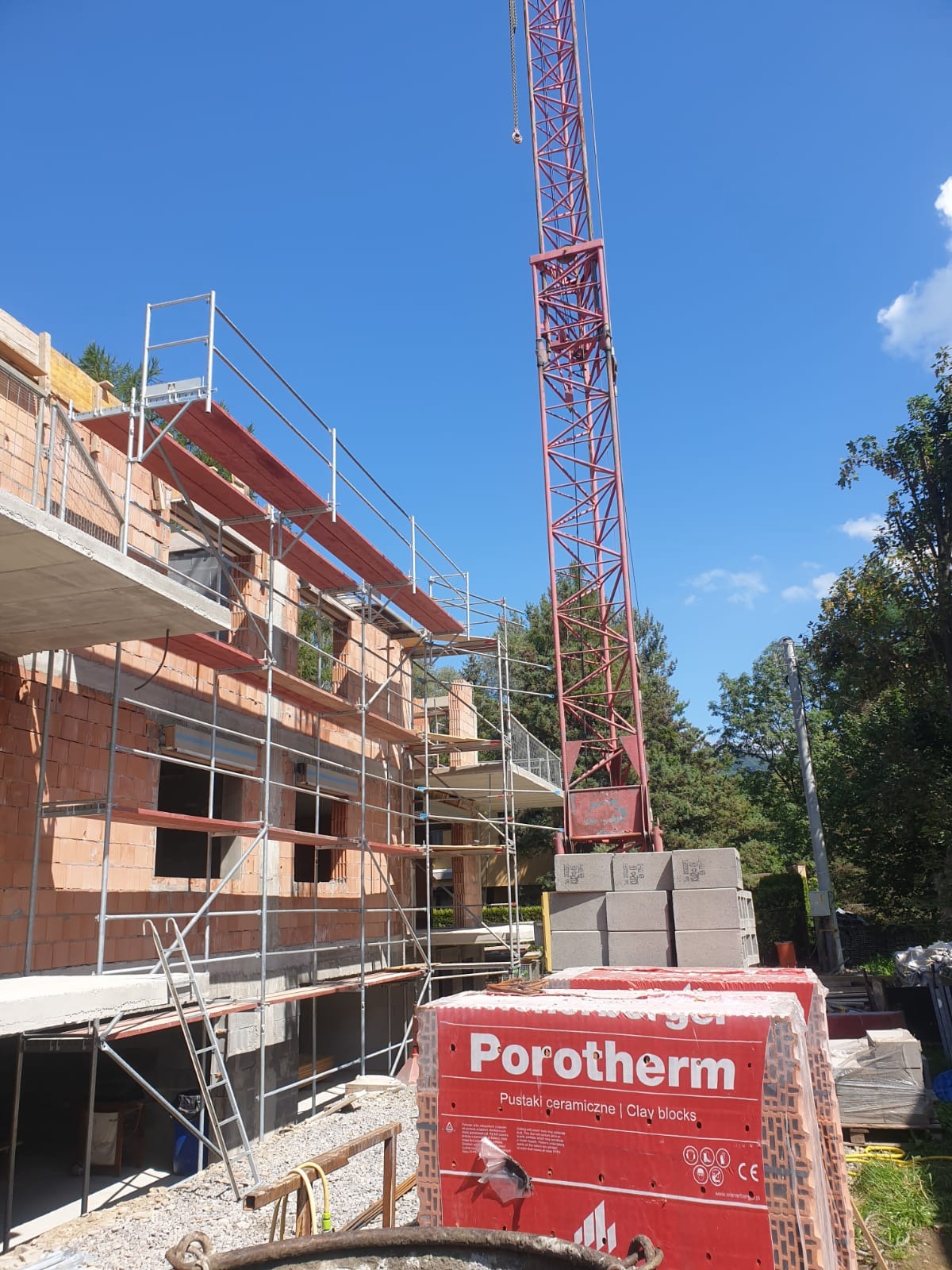 Budowa domu jednorodzinnego z czerwonej cegły Porotherm, widoczne rusztowania, dźwig i palety z materiałem budowlanym na tle błękitnego nieba.