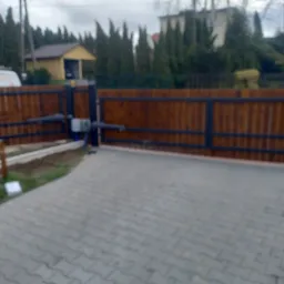 Drewniana brama wjazdowa z automatyką, widok z zewnątrz na podjazd z kostki brukowej, w tle budynki mieszkalne i wysokie drzewa.