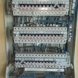 Rozdzielnica elektryczna z wieloma bezpiecznikami i przewodami, zamontowana na ścianie wykończonej panelami drewnopodobnymi, widok z bliska na szczegóły połączeń.