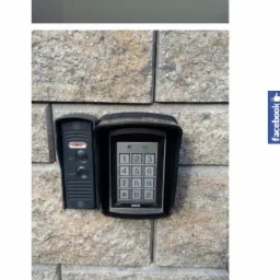 Zewnętrzny panel domofonowy z klawiaturą numeryczną i czytnikiem zbliżeniowym, zamontowany na ścianie z kamiennych bloków, nad skrzynką bezpieczników i zrzutami ekranu z monitoringu.