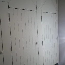 Białe, modułowe ścianki działowe z pionowymi panelami i geometrycznym wzorem na górze, widoczne zawiasy i zamki w pomieszczeniu z szarą podłogą.