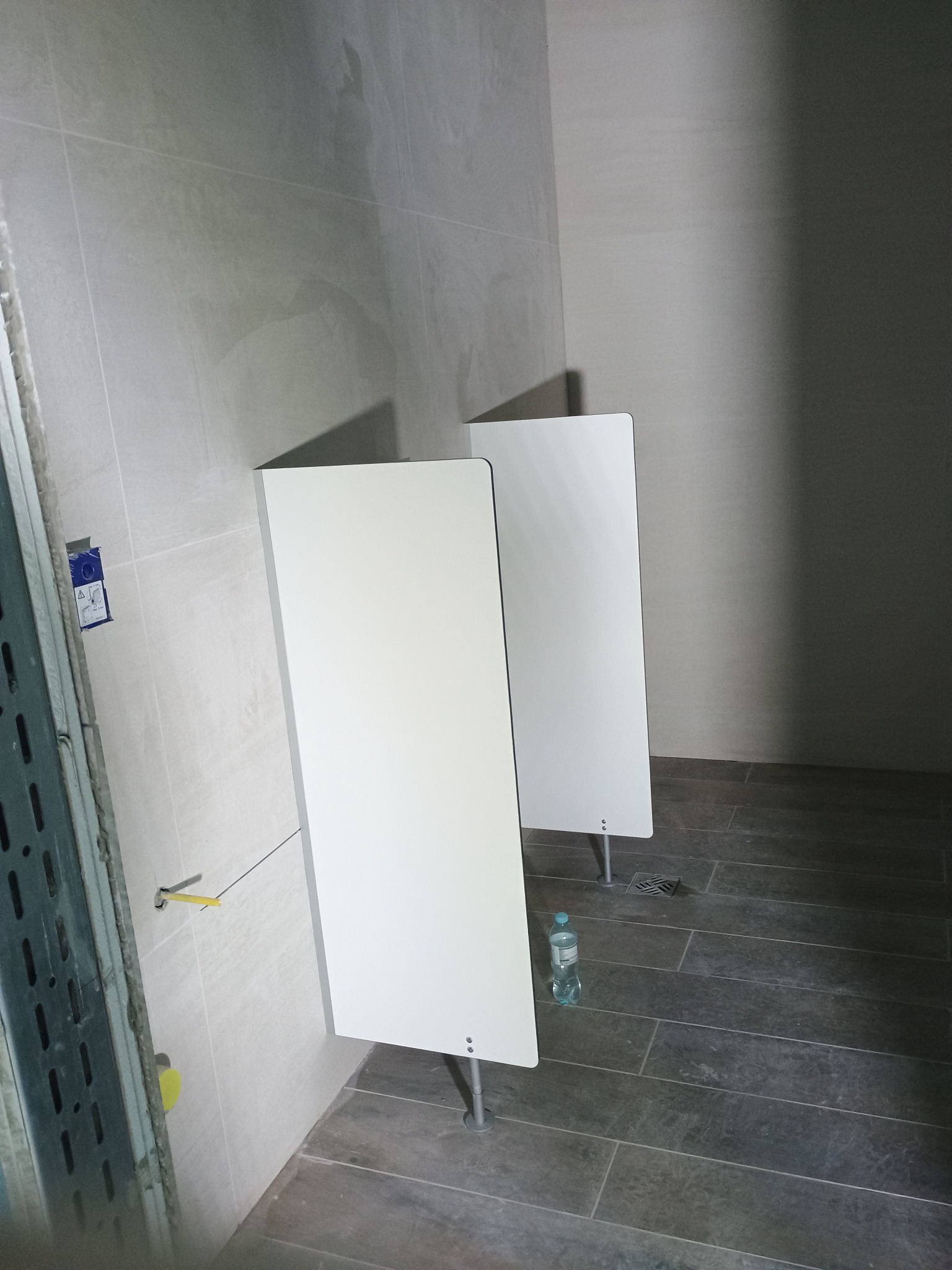 Dwa białe pisuary z przegrodami w trakcie montażu w publicznej toalecie, widoczna butelka wody na podłodze i elementy instalacji na ścianie.