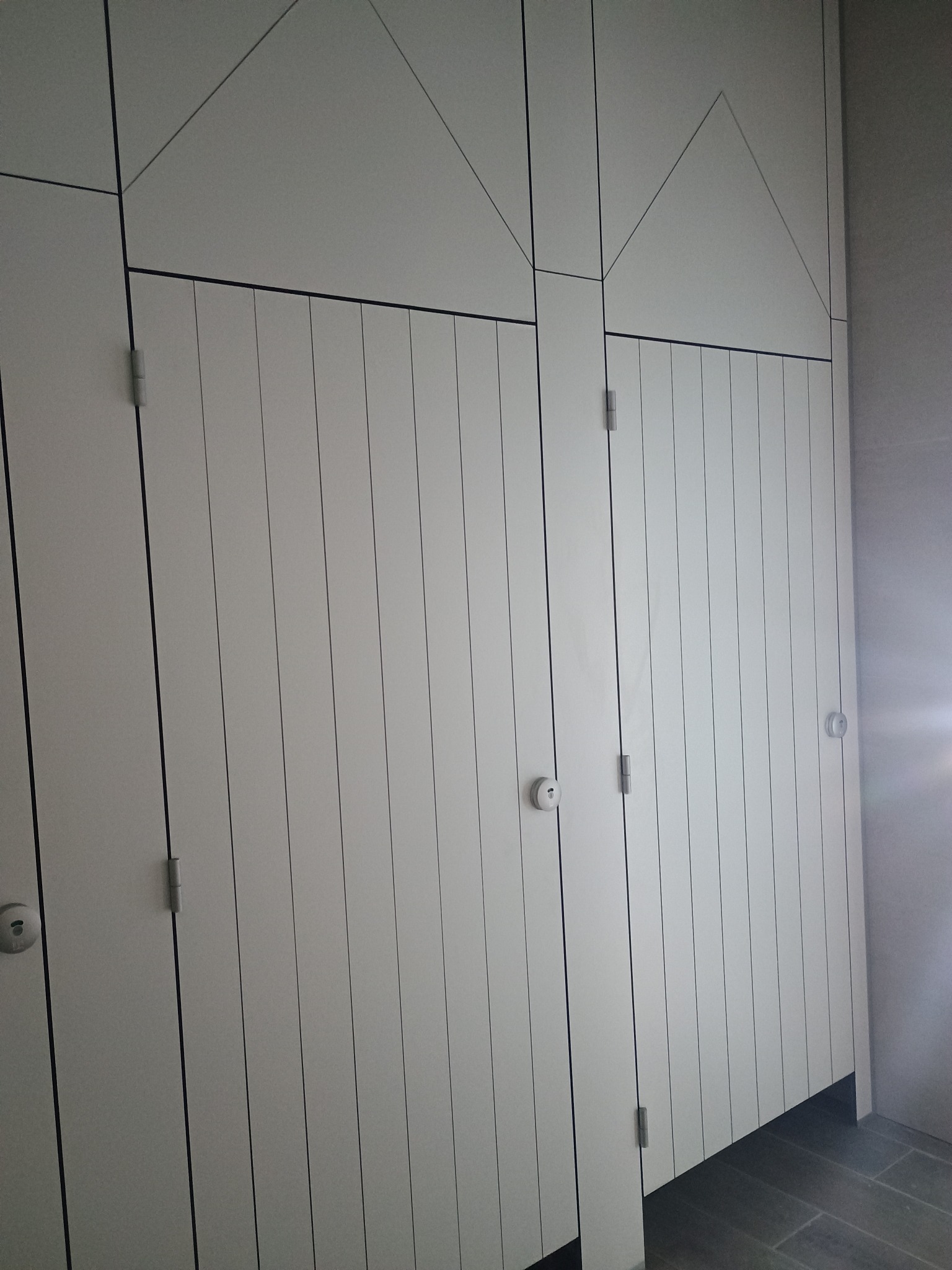 Białe, modułowe ścianki działowe z pionowymi panelami i geometrycznym wzorem na górze, widoczne zawiasy i zamki w pomieszczeniu z szarą podłogą.