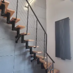 Nowoczesne schody drewniane na metalowej konstrukcji z balustradą z linek stalowych, zamontowane przy ścianie wyłożonej szarymi płytami imitującymi beton, z widocznymi gniazdkami elektrycznymi...