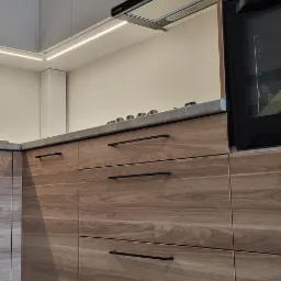 Nowoczesna zabudowa kuchenna z drewnianymi frontami szuflad, czarnymi uchwytami, szarym blatem i oświetleniem LED podszafkowym, widziana z niskiej perspektywy.