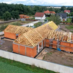 Budowa domu jednorodzinnego w stanie surowym otwartym, widok z góry na konstrukcję więźby dachowej na murach z czerwonej cegły, w tle zabudowa jednorodzinna i drzewa.