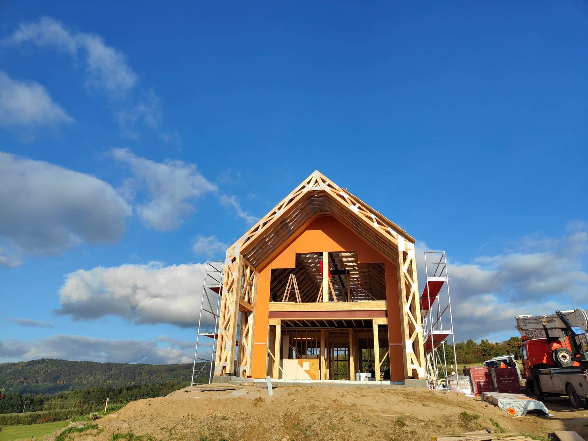 Konstrukcja domu jednorodzinnego w stanie surowym otwartym z widoczną więźbą dachową, rusztowaniami i podnośnikiem na tle błękitnego nieba z chmurami i zielonego krajobrazu.