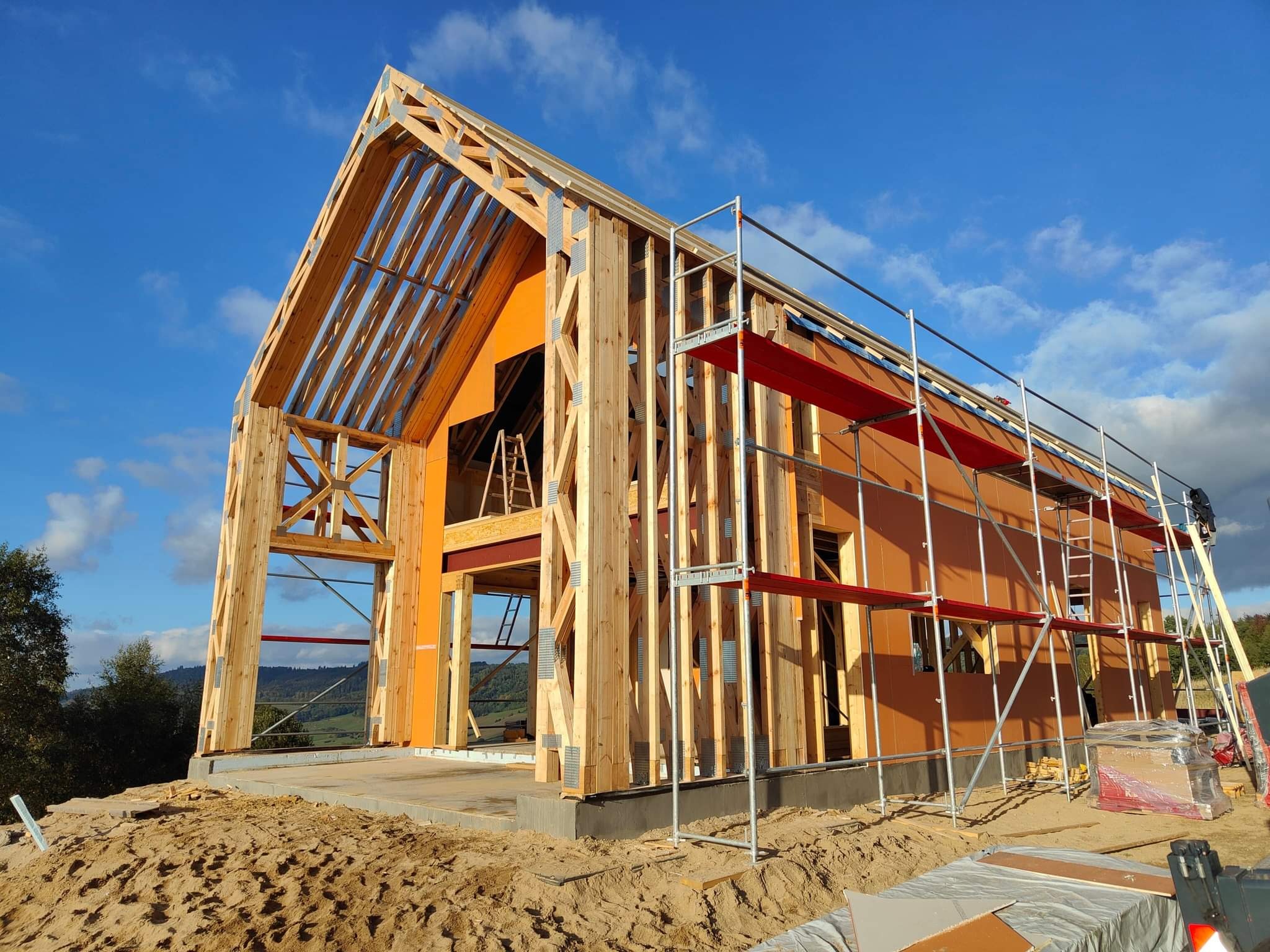 Surowa drewniana konstrukcja domu w trakcie budowy, widoczna więźba dachowa i rusztowania na tle błękitnego nieba.