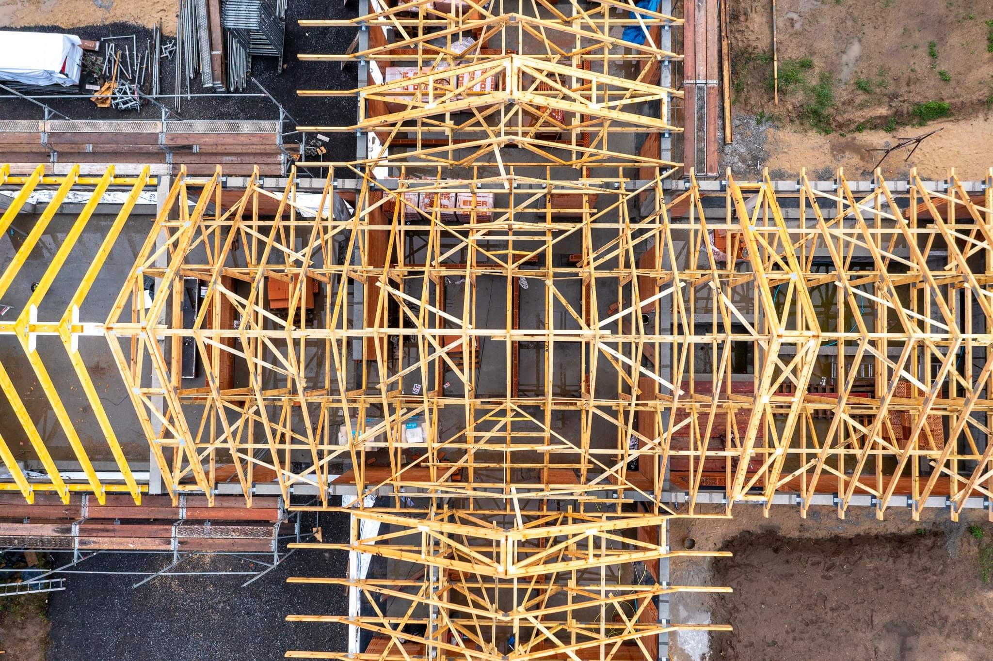 Widok z góry na konstrukcję drewnianą dachu w trakcie budowy, z widocznymi elementami deskowania i rusztowaniami w tle.
