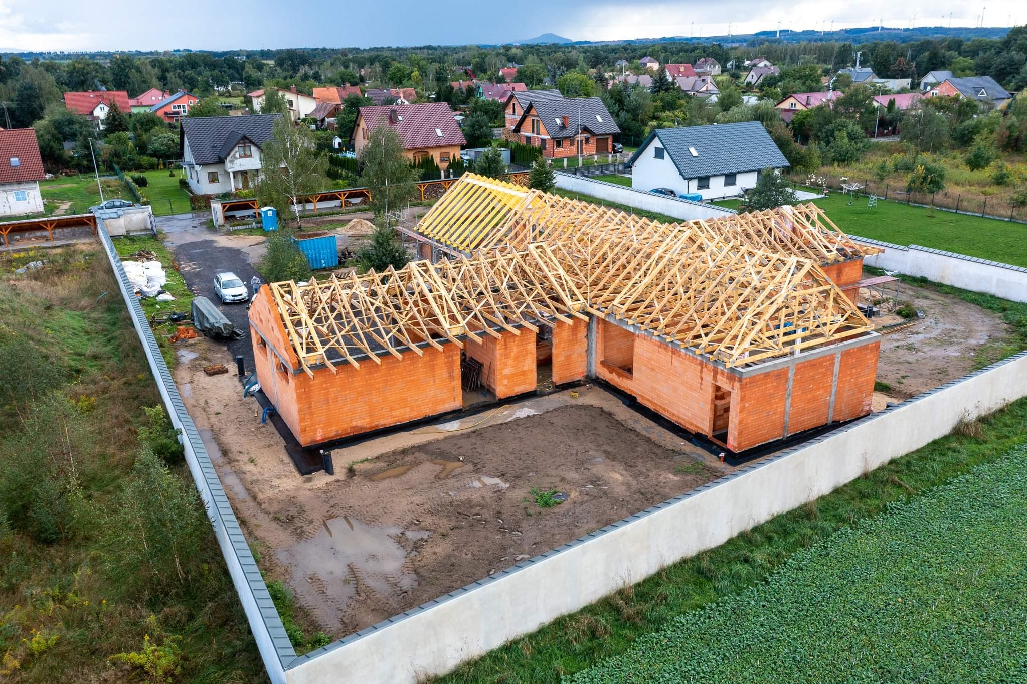 Wysoka perspektywa na budowę domu jednorodzinnego z czerwonej cegły, w trakcie montażu drewnianej konstrukcji szkieletowej dachu, otoczonego murem i zielenią, z widokiem na zabudowę mieszkalną w tle.