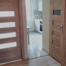 Kj-Remonty - Widok na wnętrze mieszkania po remoncie, ukazujący drewniane drzwi z przeszkleniami, podłogę z paneli oraz fragment kuchni z białymi szafkami i mozaiką nad blatem.