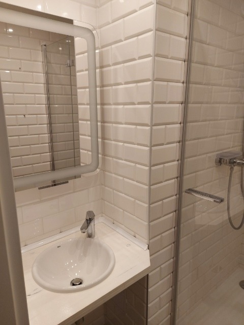 Biała umywalka z baterią, obudowana blatem, na tle ściany wyłożonej białymi, prostokątnymi płytkami imitującymi cegłę, w narożniku łazienki z kabiną prysznicową.