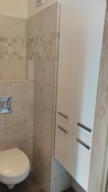 Wykończona łazienka z beżowymi płytkami, wiszącą toaletą i białą szafką z metalowymi uchwytami.