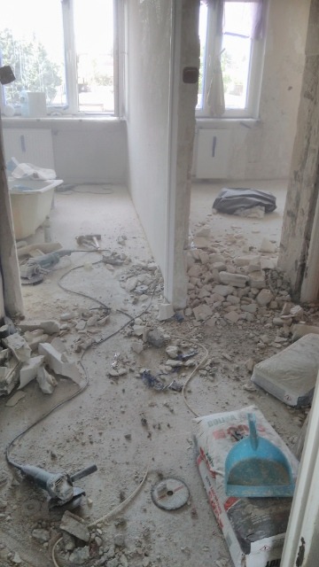 Wnętrze w trakcie demontażu: gruz, pył, narzędzia (szlifierka kątowa), worek z zaprawą oraz niebieska szufelka na podłodze; widoczne okna i fragment wanny.