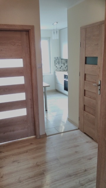 Widok na wnętrze mieszkania po remoncie, ukazujący drewniane drzwi z przeszkleniami, podłogę z paneli oraz fragment kuchni z białymi szafkami i mozaiką nad blatem.