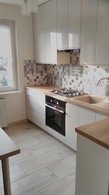 Nowoczesna kuchnia z białymi, lakierowanymi szafkami, drewnianym blatem, wzorzystą mozaiką nad blatem i płytkami podłogowymi imitującymi drewno.