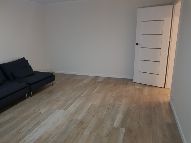 Jasne panele podłogowe ułożone w nowym mieszkaniu, widoczna biała ściana, drzwi z poziomymi frezowaniami i fragment ciemnej sofy.