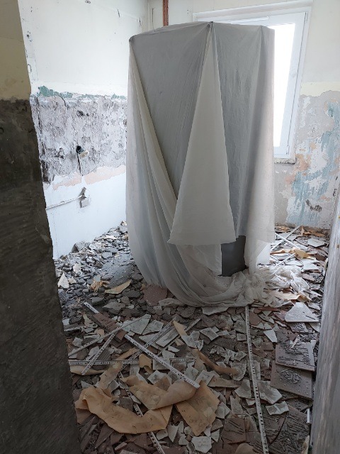 Łazienka w trakcie demontażu płytek, z gruzem na podłodze i zabezpieczoną folią wanną lub brodzikiem przy oknie.