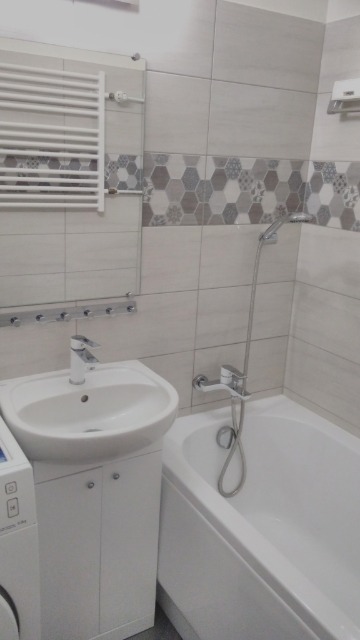 Jasna łazienka po remoncie z umywalką na szafce, wanną z prysznicem i grzejnikiem drabinkowym na ścianie wyłożonej płytkami z dekoracyjnym pasem w kształcie plastra miodu.