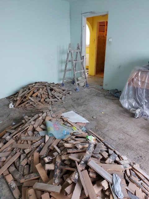 Pokój w trakcie demontażu podłogi: sterta zdemontowanych desek podłogowych, drabina malarska, widoczne ślady kleju na posadzce, ściany pomalowane na jasnozielono.