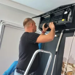 Mężczyzna na drabinie, w czarnej koszulce, instaluje czarny klimatyzator ścienny w pomieszczeniu z szarą ścianą i obrazem. Widoczne rury instalacyjne i przewody.