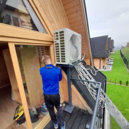 City system security - Montaż zewnętrznej jednostki klimatyzacji na wsporniku przy drewnianym domu w górach, widoczne przewody i drabina oparta o balkon.