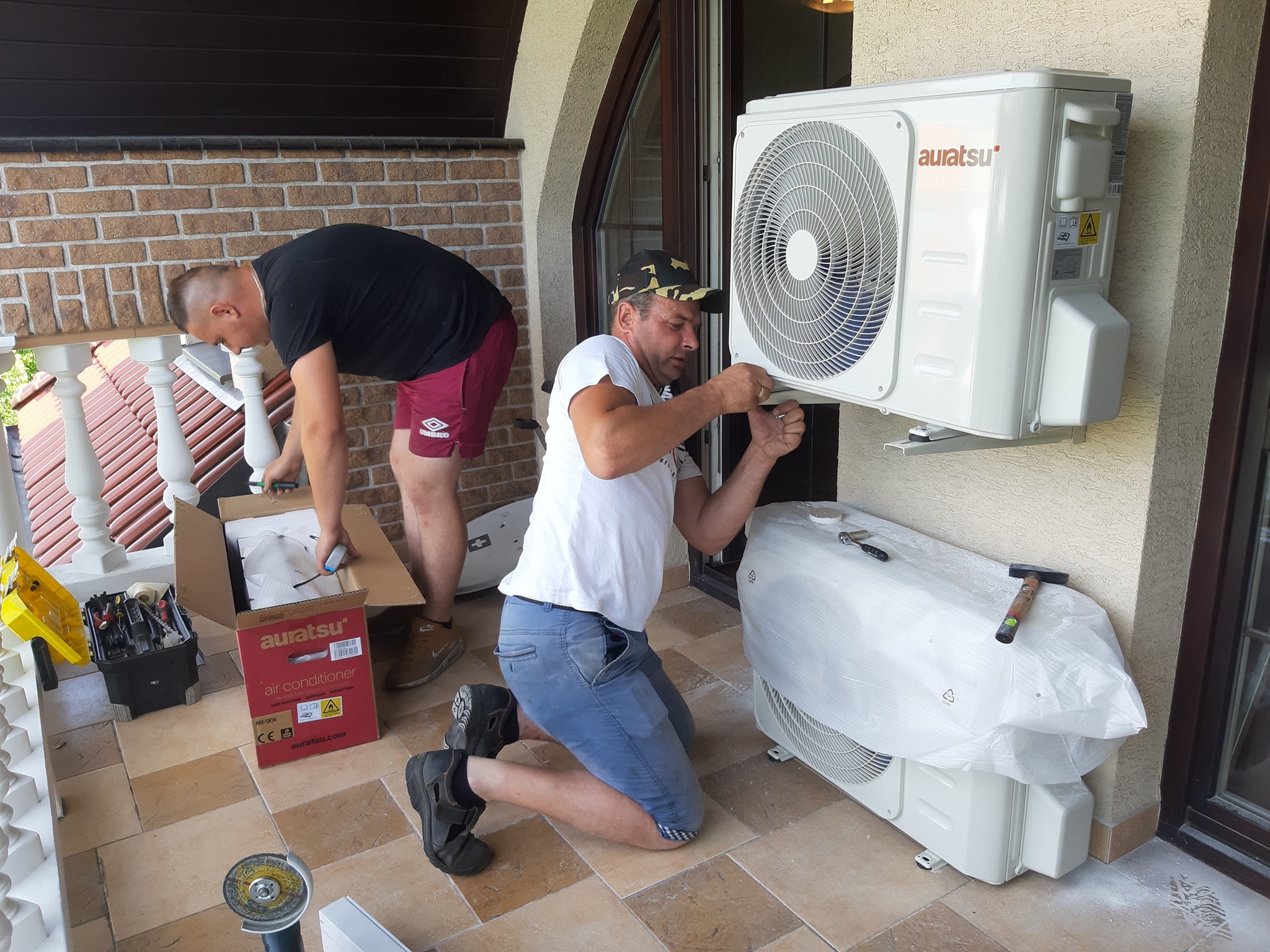 Dwóch instalatorów montuje jednostkę zewnętrzną klimatyzacji marki Auratsu na balkonie domu, jeden przykręca urządzenie do ściany, drugi rozpakowuje kolejną jednostkę z pudełka, widoczna skrzynka...