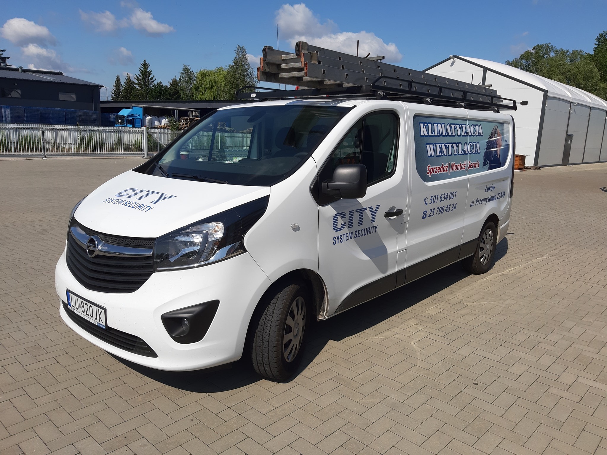 Biały samochód dostawczy firmy City System Security z drabiną na dachu, z reklamą usług klimatyzacji i wentylacji na boku, zaparkowany na brukowanym placu.