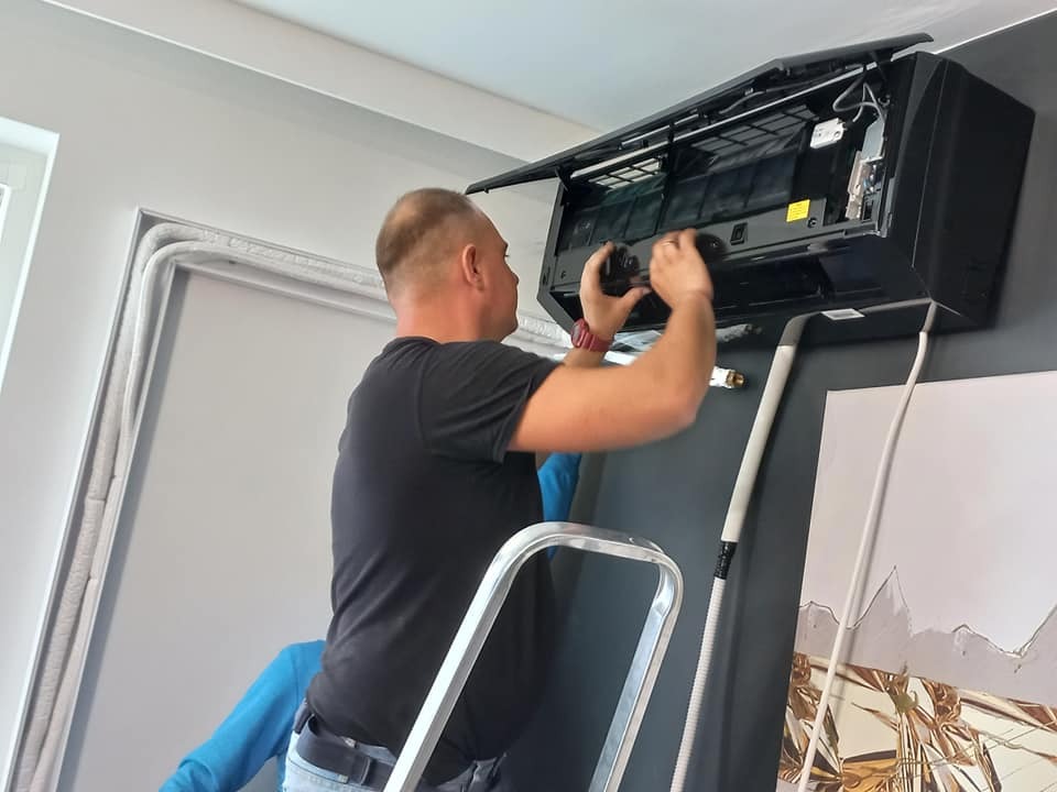 Mężczyzna na drabinie, w czarnej koszulce, instaluje czarny klimatyzator ścienny w pomieszczeniu z szarą ścianą i obrazem. Widoczne rury instalacyjne i przewody.