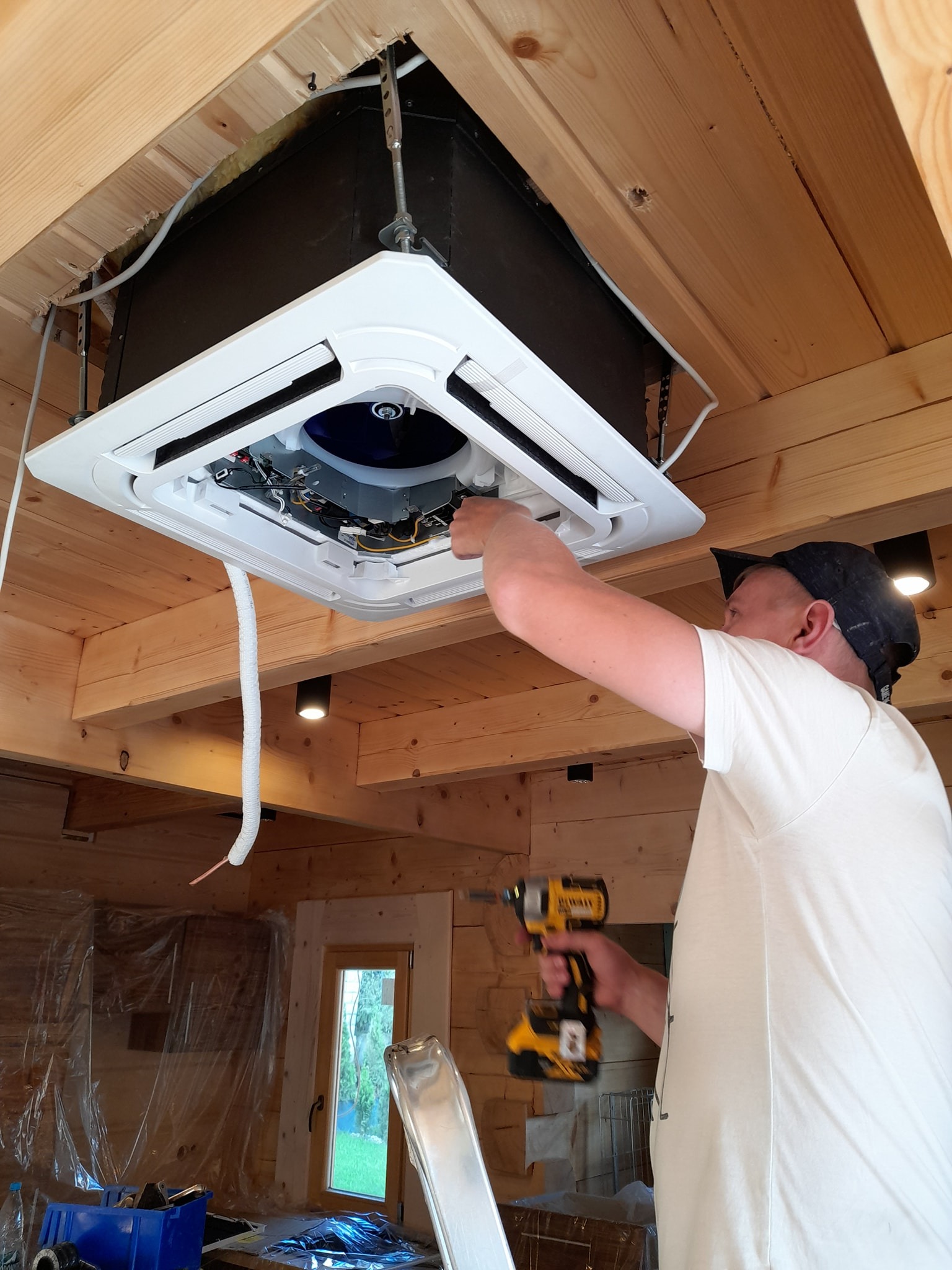 Instalacja sufitowej klimatyzacji kasetonowej w drewnianym domu, monter z wkrętarką DeWalt przy pracy, widoczne elementy montażowe i izolacja przewodów.